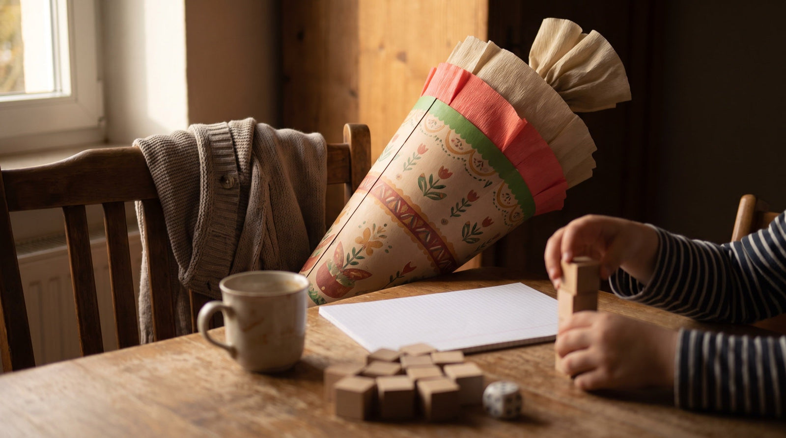 Schultüte lehnt am Küchenstuhl, davor stapeln Kinderhände Holzwürfel neben einem neuen Heft